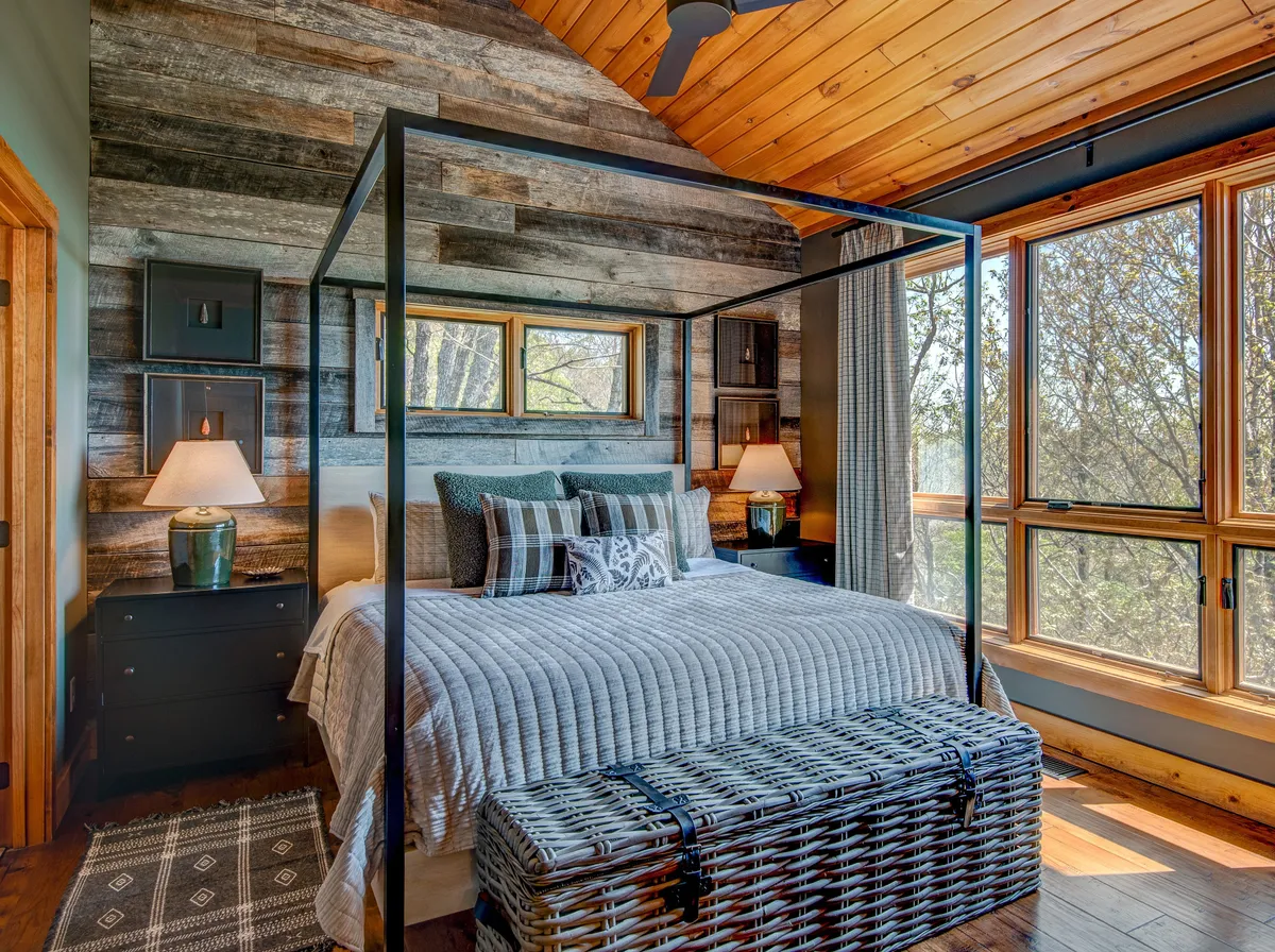 A tranquil bedroom featuring a canopy bed adorned with decorative pillows, large windows that frame scenic nature views, and a woven storage chest at the foot of the bed. Sunlight fills the room, enhancing its serene and inviting atmosphere.