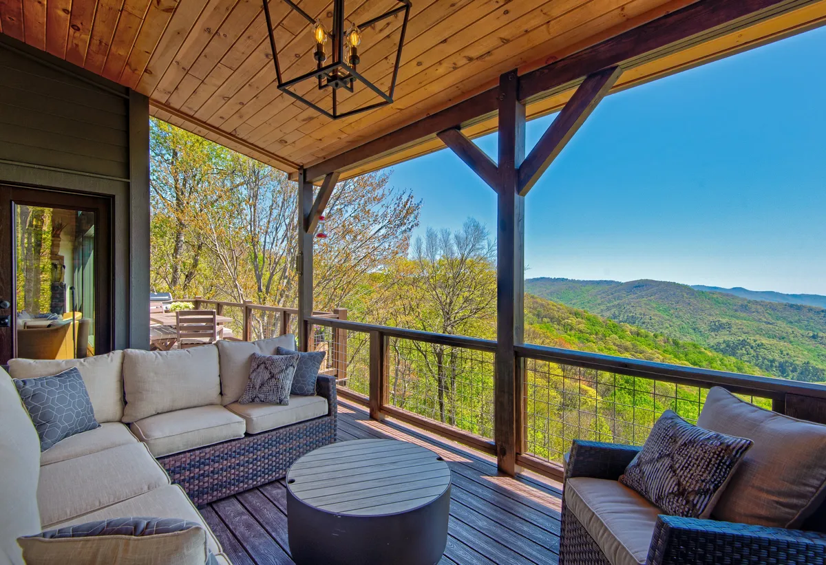 An inviting outdoor seating area with comfortable chairs and a table sits under a hanging light fixture, all positioned to take in sweeping mountain views just beyond the deck. The scene is tranquil, hinting at the perfect spot to relax, gather, or enjoy morning coffee surrounded by nature.