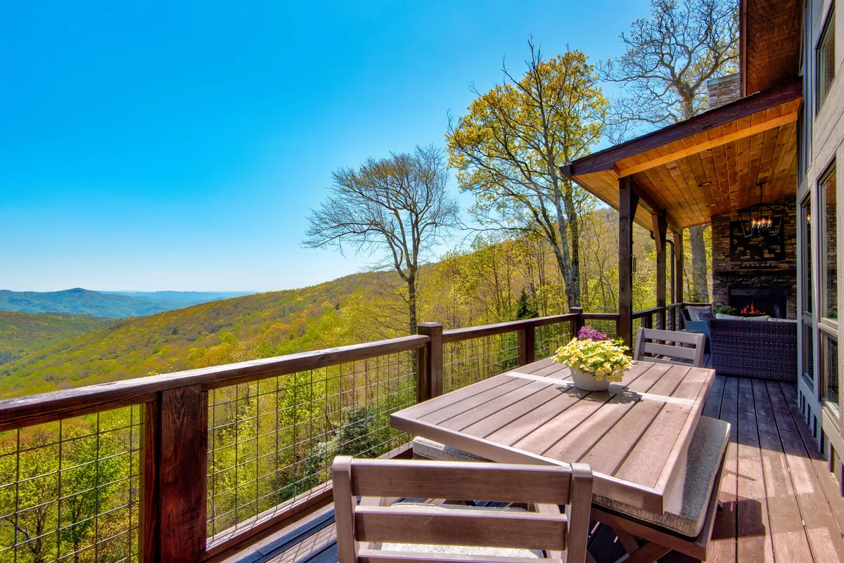 This exterior image features a dining table set on a spacious deck, surrounded by lush potted flowers and offering sweeping mountain views. The setup suggests plenty of room for relaxing or entertaining outdoors while enjoying the serenity and natural beauty of the setting.