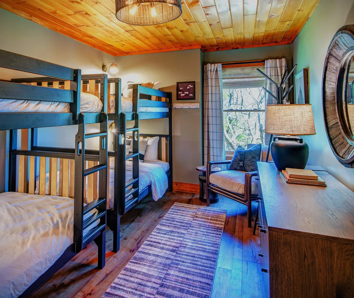 A cheerful bunk room with sturdy bunk beds, a cozy reading chair by the window, a decorative lamp, and a mirror. The room is bathed in natural light, creating an inviting space designed for children or families to unwind, play, and build memories together.