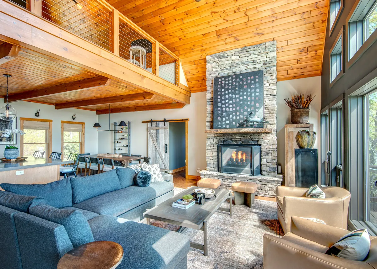 A spacious living room with a cozy fireplace, large windows offering mountain views, plush sofas, and wooden beams overhead. The setting radiates warmth and invites gathering, with the sunset visible through the windows adding to the ambiance.