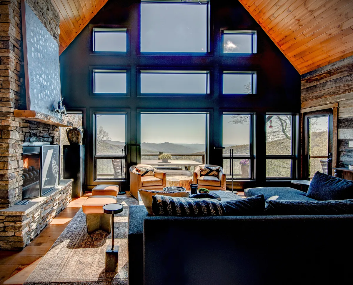 The image showcases the cabin's inviting living area, featuring expansive windows framing breathtaking mountain views, a majestic stone fireplace, and plush, comfortable seating. Artistic wall decor adds a touch of luxury, while the scene radiates warmth and tranquility—perfect for gathering with family or friends.