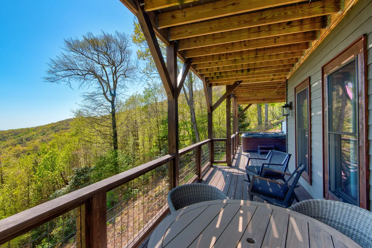 The photo features a spacious wooden deck surrounded by trees, equipped with a hot tub, a dining table, and comfortable seating. The tranquil environment offers both relaxation and opportunities for outdoor dining or socializing.