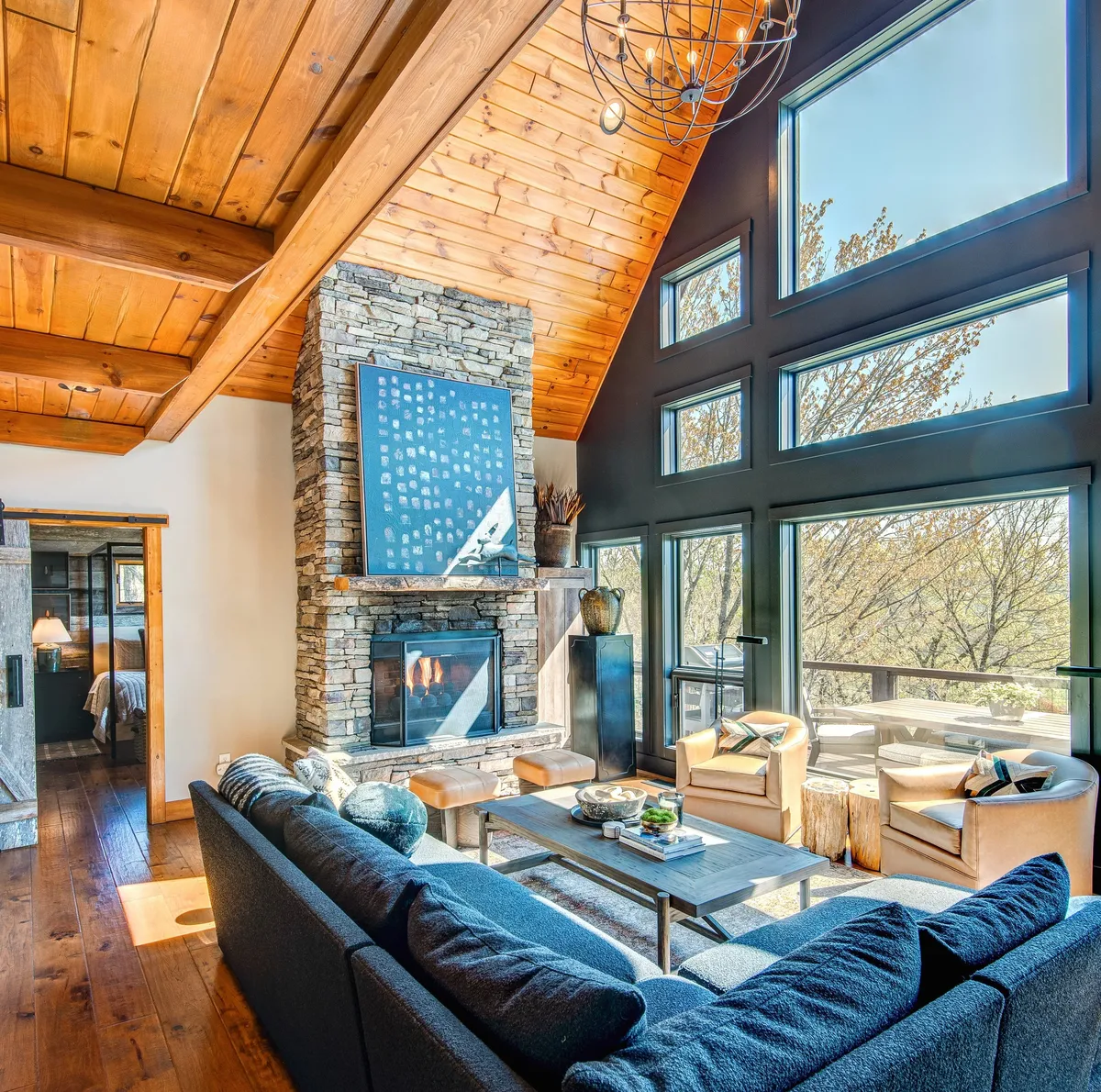 A bright, inviting living room featuring a large stone fireplace and floor-to-ceiling windows that showcase breathtaking mountain views. The space is anchored by comfortable seating and a stylish coffee table, and the wooden ceiling adds a cozy, rustic touch.