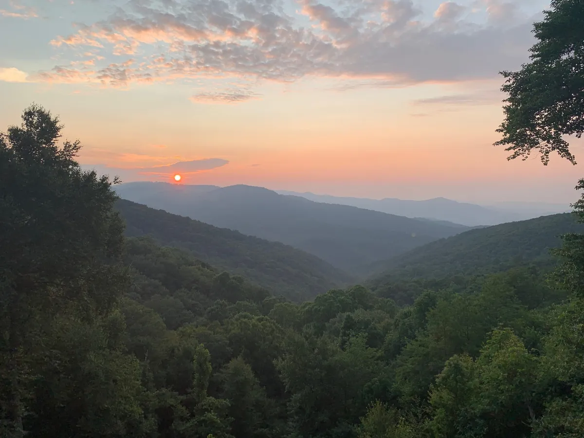 A panoramic view of the mountains at sunset, rolling hills covered in lush greenery, and tranquil skies. The landscape evokes the peace and beauty of the Blue Ridge Parkway and nearby hiking trails.