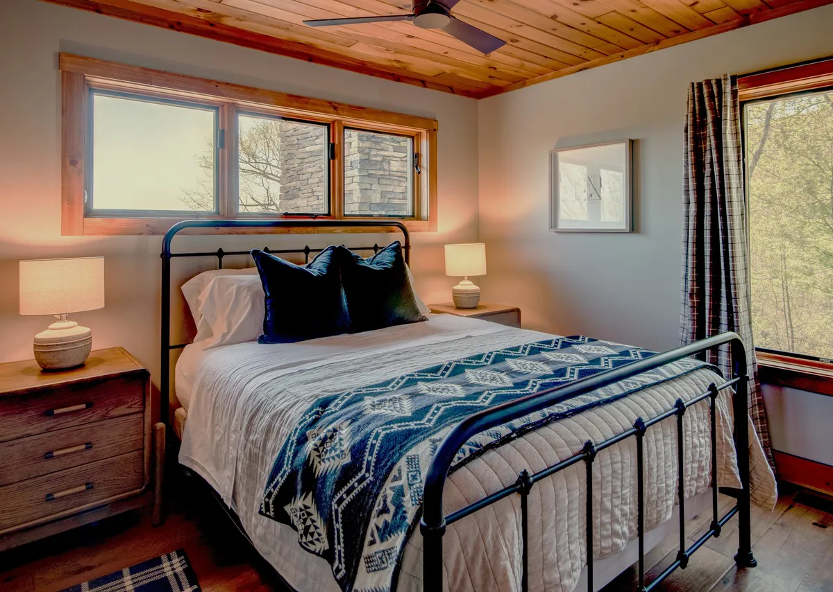 A cozy bedroom with a comfortable bed dressed in a patterned throw, nightstands with glowing lamps, and windows flooded with natural light. The simple yet inviting decor enhances the sense of comfort and privacy.