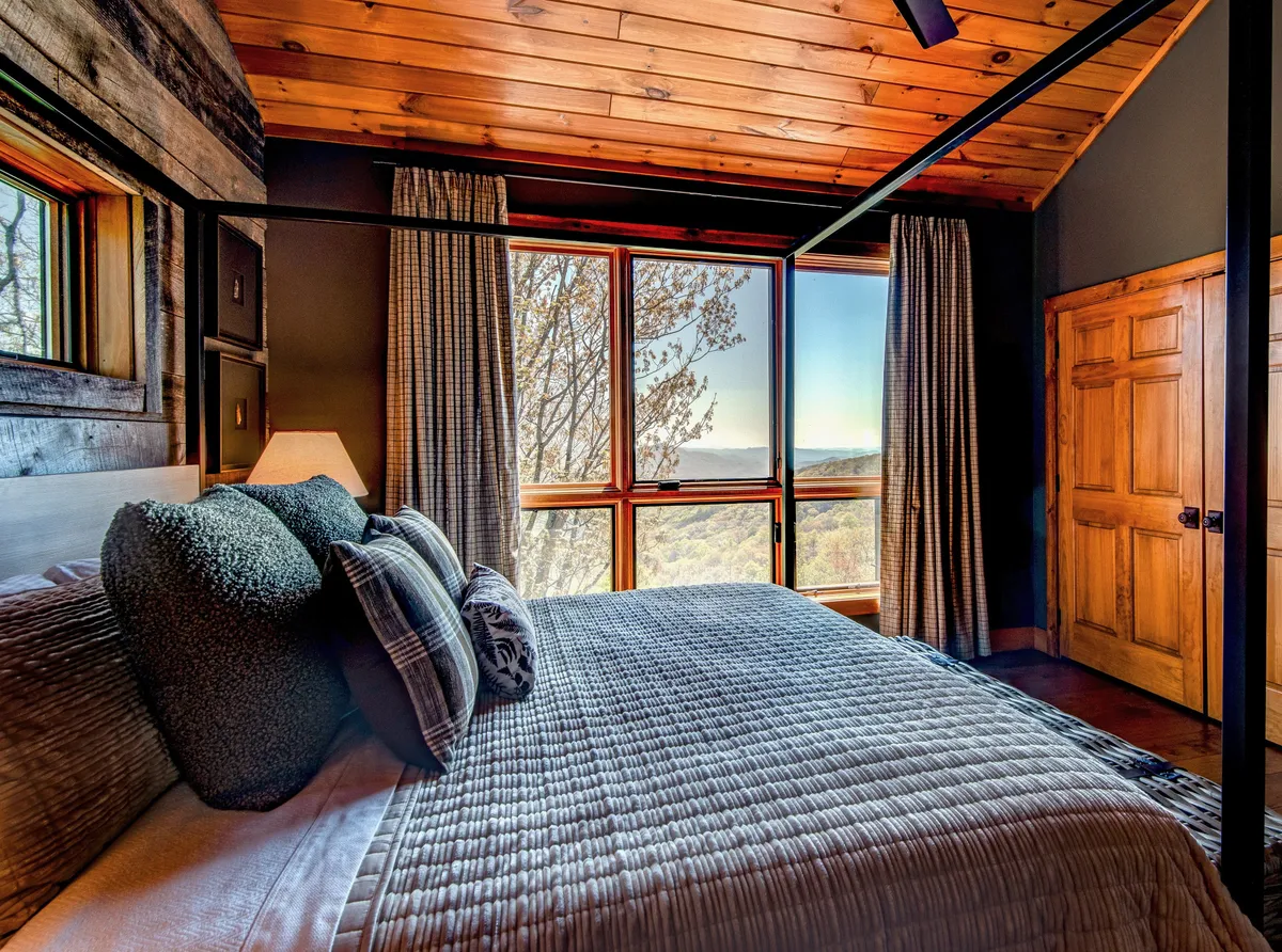 A tranquil bedroom featuring a canopy bed and cozy bedding, oriented toward oversized windows that reveal stunning mountain views. The natural light pouring in emphasizes the close relationship between the home and its scenic surroundings.