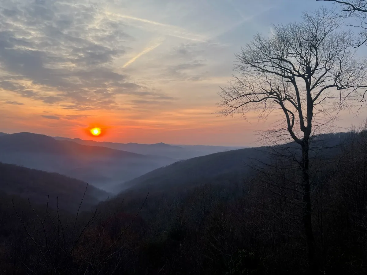 A scenic landscape at sunset, highlighting a dramatic mountain range and silhouetted trees. The photo captures the region's natural beauty, inviting guests to explore Blowing Rock, the Blue Ridge Parkway, and appreciate the picturesque environment.