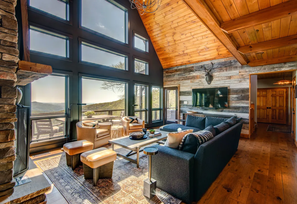 This image features a cozy seating area arranged around a stone fireplace, with expansive windows revealing sweeping mountain views beyond. The rustic wooden accents and comfortable furnishings evoke the charm of mountain living, while the view through the glass connects the interior to the wild landscape outside.